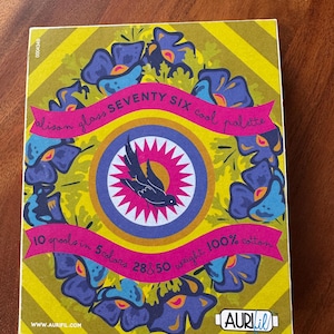 May include: A rectangular product box for Alison Glass Seventy Six cool palette thread, featuring a yellow and green striped background with a central purple circle, a black bird illustration, and blue floral motifs. Text includes 10 spools in 5 colors, 28 & 50 weight, 100% cotton, and Aurifil branding.