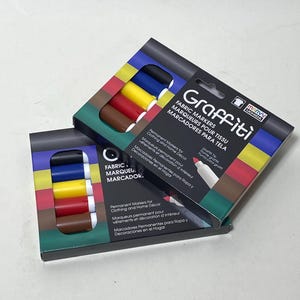 May include: Two boxes of Graffiti fabric markers. Each box contains six markers in various colors: black, blue, yellow, red, and brown. The packaging includes the text "FABRIC MARKERS" and "Permanent Markers for Clothing and Home Décor."