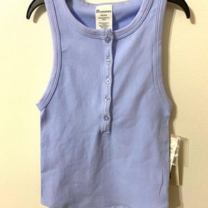May include: A light purple tank top with a henley neckline and a row of buttons down the front. The top is hanging on a black hanger. The tag reads "No Boundaries".
