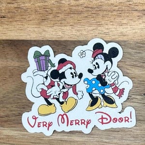 May include: A die-cut sticker featuring Mickey Mouse in a red Santa suit holding a purple gift and Minnie Mouse in a blue polka-dot dress and red scarf. The text "Very Merry Door!" is printed in red at the bottom of the white-bordered sticker.