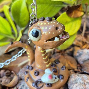 May include: A handmade brown snake ornament with a detailed design. The snake has a light brown body with dark brown spots and a white and pink mouse. The snake has a chain attached to it. The background is blurred with green leaves and rocks.
