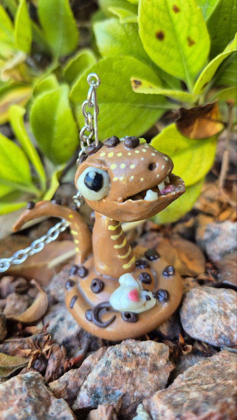 May include: A handmade brown snake ornament with a detailed design. The snake has a light brown body with dark brown spots and a white and pink mouse. The snake has a chain attached to it. The background is blurred with green leaves and rocks.