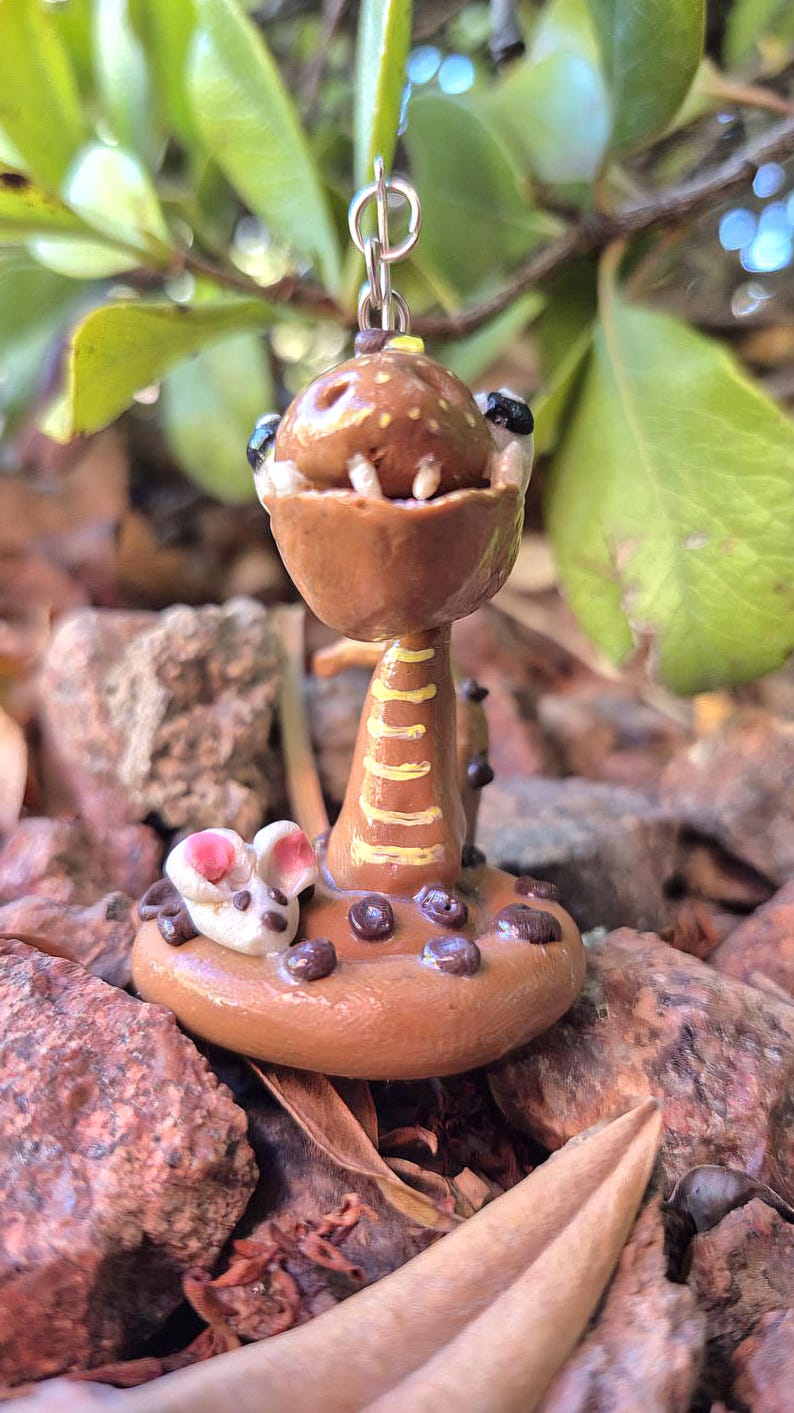 May include: A handmade polymer clay pendant depicting a brown, cartoonish snake with an open mouth and small white teeth. The pendant has a yellow and brown stem and a small mouse at the base. It hangs from a silver-coloured chain.