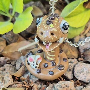 May include: A handmade brown snake pendant with black spots and a small mouse charm. The snake has large eyes, a pink tongue, and is on a silver chain. The pendant is set against a background of rocks and leaves.