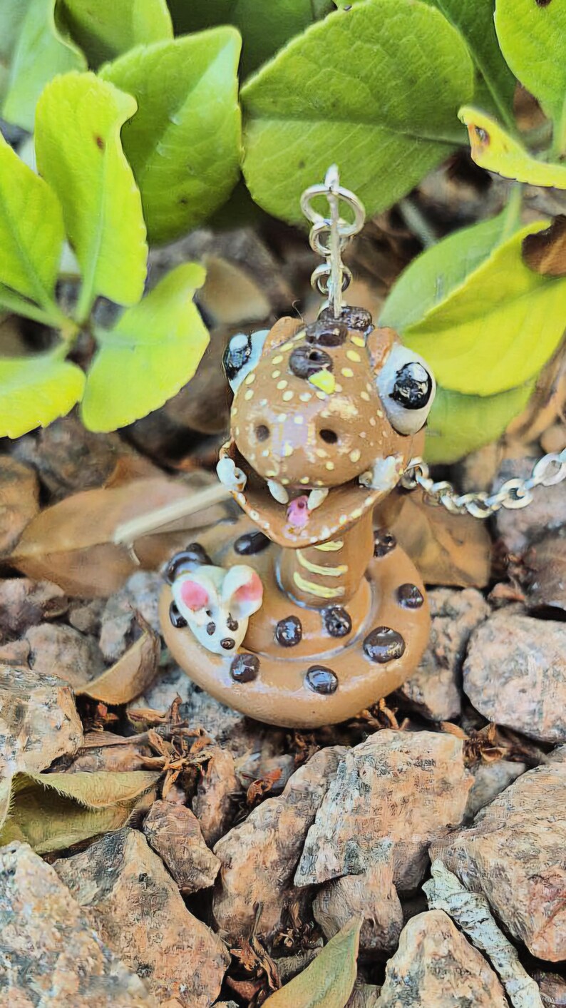 May include: A handmade brown snake pendant with black spots and a small mouse charm. The snake has large eyes, a pink tongue, and is on a silver chain. The pendant is set against a background of rocks and leaves.
