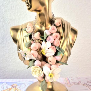 Op de afbeelding: Een gouden buste sculptuur versierd met een ketting met roze kraalachtige elementen, witte bloemen en groene bladeren. De ketting is om de nek van de buste gedrapeerd, waardoor een decoratieve weergave ontstaat. De buste staat tegen een zachte achtergrond.