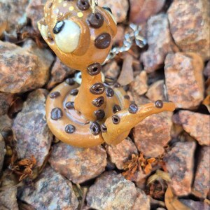 May include: A handmade snake pendant in a whimsical style. The snake is light brown with dark brown spots and a white face. It is coiled on a bed of small rocks. The pendant is attached to a silver chain.