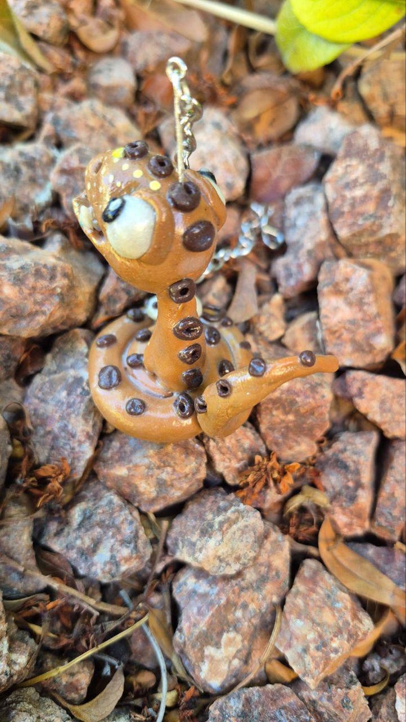 May include: A handmade snake pendant in a whimsical style. The snake is light brown with dark brown spots and a white face. It is coiled on a bed of small rocks. The pendant is attached to a silver chain.
