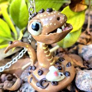 May include: A handmade brown snake figurine with white spots and black detailing. The snake has a smiling face, visible teeth, and a small white mouse figurine attached. A silver chain is attached to the snake's head. The figurine rests on a bed of rocks.