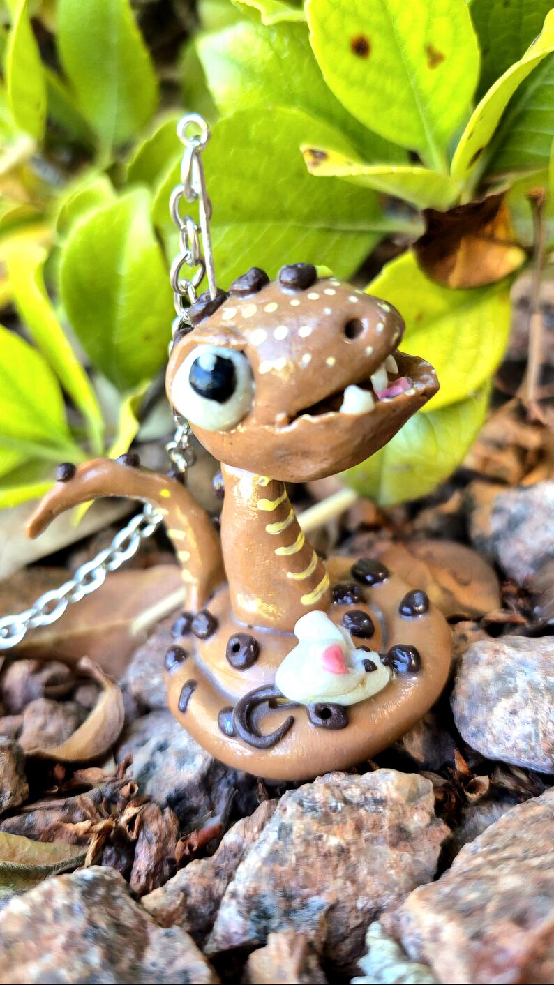 May include: A handmade brown snake figurine with white spots and black detailing. The snake has a smiling face, visible teeth, and a small white mouse figurine attached. A silver chain is attached to the snake's head. The figurine rests on a bed of rocks.