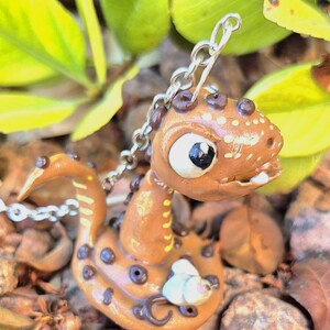 May include: A handcrafted, brown polymer clay dragon pendant with a silver chain. The dragon has white and black eyes, dark brown spots, and a small white flower accent. The background features green leaves and small rocks.