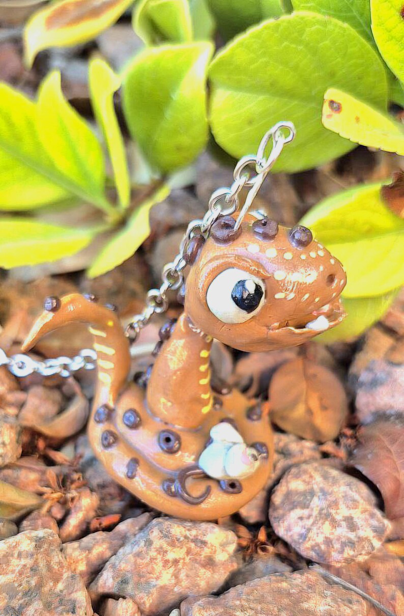 May include: A handcrafted, brown polymer clay dragon pendant with a silver chain. The dragon has white and black eyes, dark brown spots, and a small white flower accent. The background features green leaves and small rocks.
