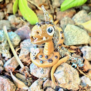 Peut inclure: Une figurine de serpent faite à la main, de couleur beige, avec des taches brun foncé et de grands yeux bleus. Le serpent a un petit compagnon souris et est attaché à une chaîne argentée, probablement un porte-clés ou un pendentif. Le fond présente des roches et du feuillage.