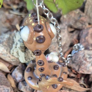 May include: A brown snake-shaped keyring with a silver chain. The snake has dark brown spots and large white eyes. The head has yellow spots. The keyring is made of clay and is a unique accessory.