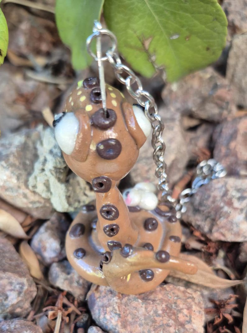 May include: A brown snake-shaped keyring with a silver chain. The snake has dark brown spots and large white eyes. The head has yellow spots. The keyring is made of clay and is a unique accessory.