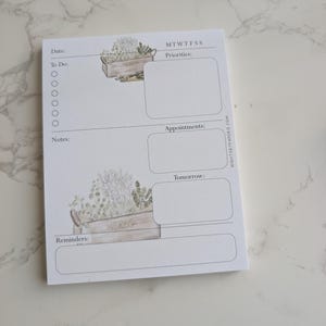 May include: A white notepad with a botanical design featuring a wooden crate filled with herbs. The notepad includes sections for date, to-do list, priorities, appointments, notes, reminders, and the days of the week.