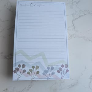 Könnte beinhalten: Ein rechteckiger Notizblock mit linierten Seiten und dem Wort "notes" in Schreibschrift oben. Der untere Teil zeigt ein farbenfrohes Zickzack-Design und florale Illustrationen. Die Website-Adresse nightskypaperie.com ist unten rechts aufgedruckt.