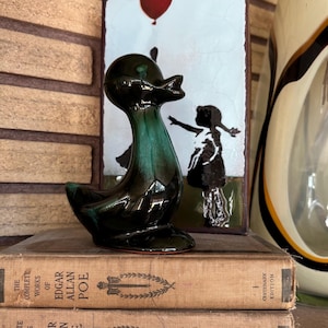 May include: A glossy dark green ceramic duck figurine positioned in front of a rectangular art piece depicting a silhouette of a child reaching for a red balloon. The duck rests on two stacked books titled The Complete Works of Edgar Allan Poe.