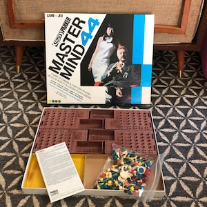 May include: A vintage Parker Brothers Master Mind 44 game in its original box. The box features a blue and white design with a photo of three people. The game board is brown and has a grid of holes. The game includes coloured pegs and a plastic bag of game pieces.