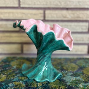 May include: A ceramic pedestal bowl featuring a ruffled, flared rim. The interior of the bowl is a solid light pink, while the exterior and the base are finished in a glossy, marbled dark green glaze with a smooth, undulating, wave-like form.