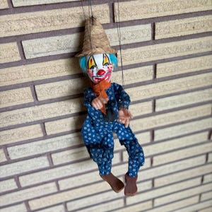 May include: A marionette clown puppet featuring a straw hat, bright blue hair, and a painted face with red accents. The puppet wears a blue jumpsuit with a small white floral pattern, an orange neck ruffle, and brown shoes, suspended by black strings.