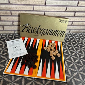 May include: Backgammon board game set with a gold-coloured box and a board featuring black, white, and orange triangles. Includes dark brown and light brown game pieces, and a printed instruction sheet. The box reads "Backgammon".