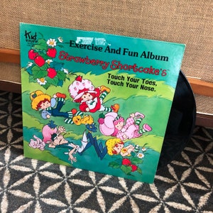 May include: A vintage vinyl record album titled "Exercise And Fun Album Strawberry Shortcake's". The cover features cartoon illustrations of Strawberry Shortcake characters in a green field with strawberries and flowers. The text "Touch Your Toes, Touch Your Nose" is also visible.