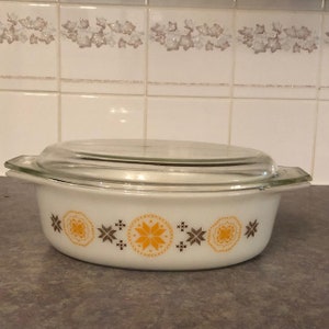 May include: An oval white casserole dish with a clear glass lid. The dish has a decorative band of orange and brown starburst and floral designs. The dish is designed for baking and serving.