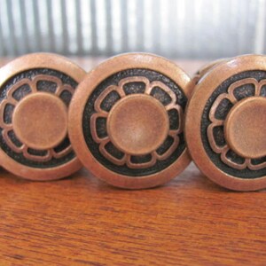 May include: A close-up of a row of copper-coloured drawer pulls with a black floral design. The pulls are round and have a raised centre.