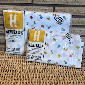 May include: Two packs of Heritage brand no-iron sheets and pillowcases. The sheets are white with a floral print of pink, yellow, and blue flowers. The packages are labelled with the brand name, product type, and size. The sheet pack is labelled "Double Flat Sheet" and measures 81" x 100". The pillowcase pack is labelled "Two Pillowcases" and measures 21" x 33".