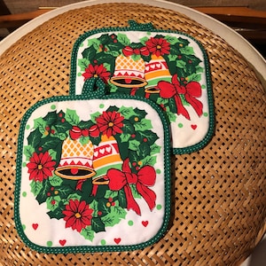 May include: Two green and white Christmas pot holders with a wreath design and red bows. The wreath has holly leaves and red flowers. There are two gold bells with red and white ribbons on each pot holder.