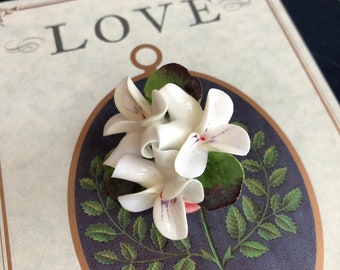 May include: A white porcelain flower brooch with a green stem and leaves. The flower has a delicate, ruffled design and a hint of purple in the center. The brooch is set against a background with the word "LOVE" printed on it.