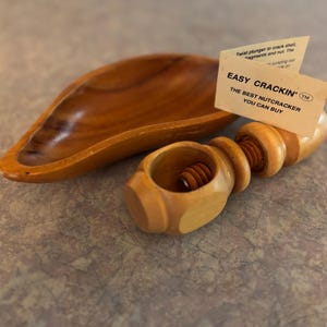 May include: A wooden nutcracker with a screw mechanism and a matching wooden bowl. The nutcracker has the text "EASY CRACKIN' THE BEST NUTCRACKER YOU CAN BUY" on a small tag. The bowl is oval-shaped.