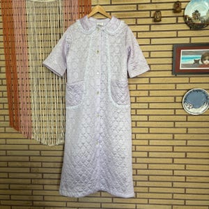 May include: A lavender quilted dressing gown with white lace trim and a button-up front. The gown has two pockets and a collar.