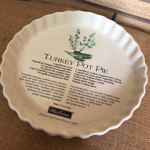 May include: A white ceramic pie dish with a recipe for Turkey Pot Pie. The recipe includes ingredients such as chicken soup base, water, celery, onion, evaporated skimmed milk, plain flour, cooked turkey breast, frozen mixed vegetables, Watkins Poultry Seasoning, black pepper, and phyllo pastry. The dish is decorated with a green and white illustration of herbs.