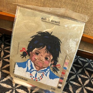 May include: A needlepoint kit featuring a girl with dark hair and a blue shirt. The kit includes a beige canvas, yarn, and instructions. The kit is made in Denmark and is 100% cotton. The kit is labelled "ARTISANA".