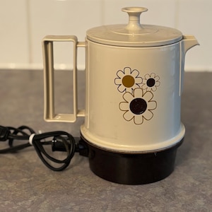 Peut inclure: Cafetière électrique crème avec une base et une poignée marron. Le pot présente un motif floral marron et or. Un cordon d'alimentation noir est attaché. La cafetière est conçue pour préparer du café.