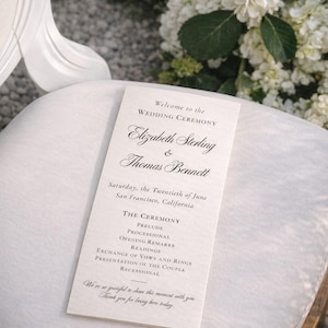 Può includere: Un programma della cerimonia nuziale su un cuscino bianco. Il programma elenca i nomi di Elizabeth Sterling e Thomas Bennett, la data e i dettagli della cerimonia. Fiori bianchi e una candela sullo sfondo.