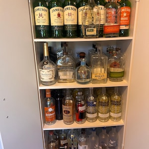 May include: A white cabinet filled with various glass liquor bottles arranged on four shelves. The collection includes multiple Jameson Irish Whiskey bottles, Bulleit Bourbon, Hennessy, Woodford Reserve, Mr. Black, Tito's Vodka, Absolut Vodka, and Grey Goose.