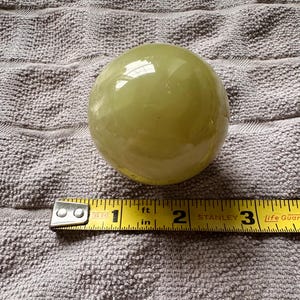 May include: A pale green, spherical object, approximately 2.5 inches in diameter, sits on a textured surface. A yellow measuring tape is placed next to the sphere, indicating its size. The sphere has a glossy finish.