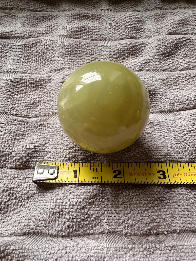 May include: A pale green, spherical object, approximately 2.5 inches in diameter, sits on a textured surface. A yellow measuring tape is placed next to the sphere, indicating its size. The sphere has a glossy finish.