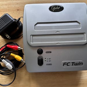 May include: A silver Yobo Gameware FC Twin video game console featuring two cartridge slots, a reset button, a power switch, and indicator lights for 8-bit and 16-bit modes. Included are a black power adapter and a set of red, yellow, and white RCA cables.