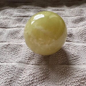 May include: A smooth, spherical, pale yellow object with a glossy surface. The sphere is resting on a textured, gray surface. The object reflects light, creating highlights and shadows.