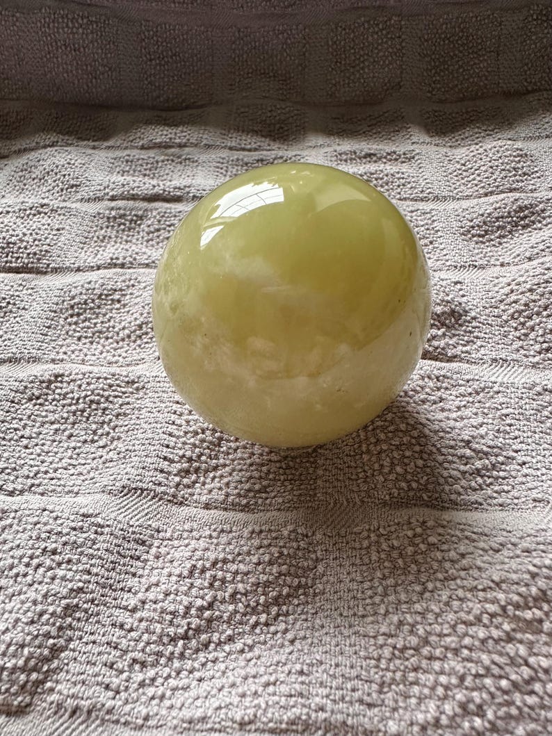 May include: A smooth, spherical, pale yellow object with a glossy surface. The sphere is resting on a textured, gray surface. The object reflects light, creating highlights and shadows.
