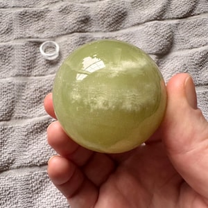 May include: A light green, translucent sphere, possibly a crystal ball, held in a hand. The sphere has a smooth surface with some internal clouding. A small, clear ring sits nearby on a textured, gray surface.
