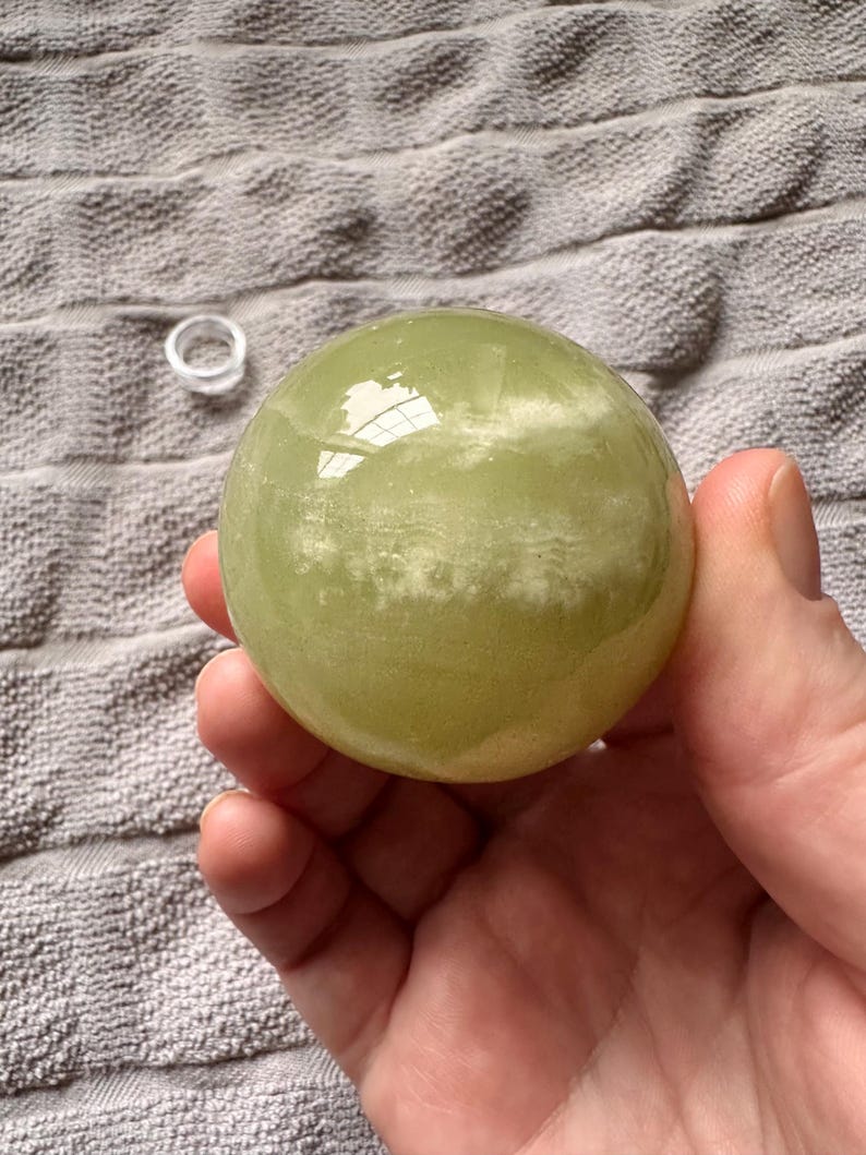 May include: A light green, translucent sphere, possibly a crystal ball, held in a hand. The sphere has a smooth surface with some internal clouding. A small, clear ring sits nearby on a textured, gray surface.