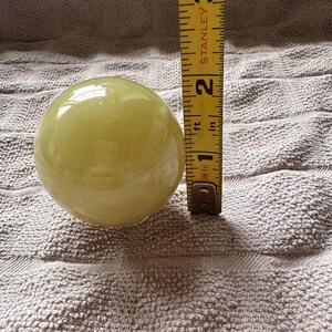 May include: A pale yellow, spherical object, approximately 2 inches in diameter, is positioned next to a yellow measuring tape. The tape shows measurements in inches and feet. The object rests on a textured, light gray surface.