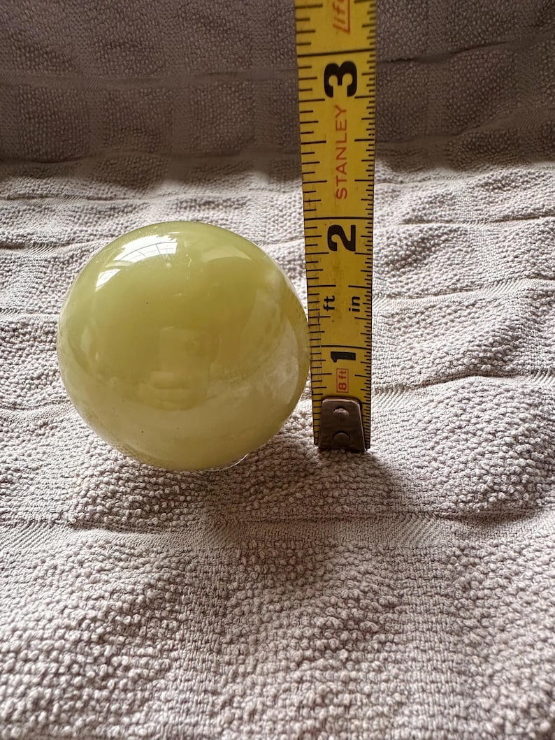 May include: A pale yellow, spherical object, approximately 2 inches in diameter, is positioned next to a yellow measuring tape. The tape shows measurements in inches and feet. The object rests on a textured, light gray surface.