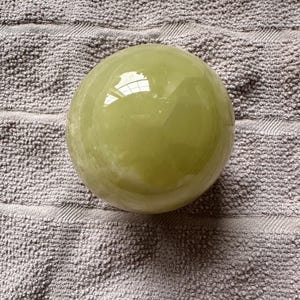 May include: A smooth, spherical, pale green object with a glossy finish. The object is resting on a textured, gray surface. The sphere reflects light, creating highlights on its surface.