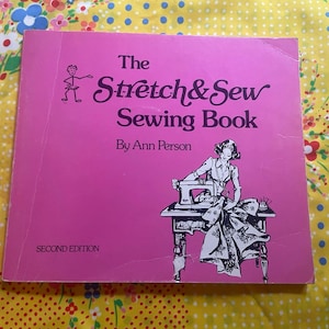 May include: A pink softcover book titled The Stretch & Sew Sewing Book by Ann Person, Second Edition. The cover features a dark purple illustration of a person using a sewing machine at a table and a small stick figure logo in the top left corner.